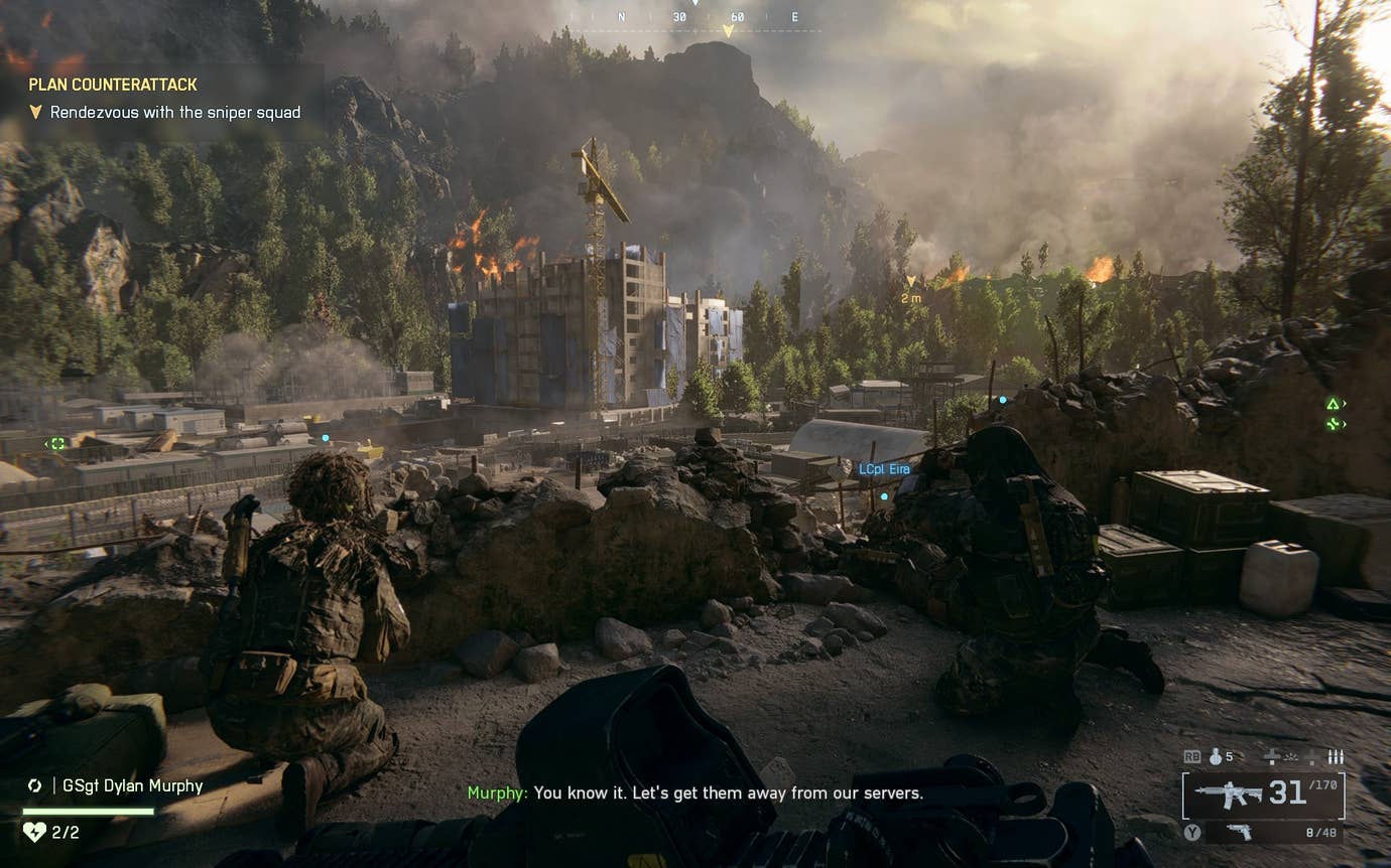 A view of soldiers grouped behind low rocks on a hillside looking down at a big concrete building in a forested valley, from Battlefield 6.